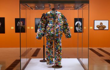 Joe Lycett's colourful suit made of numerous small teddy bears plastic displayed in a glass case in art gallery.