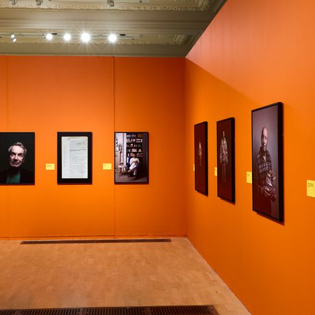 Portraits photographs showcased on bright orange walls in a modern art gallery setting.