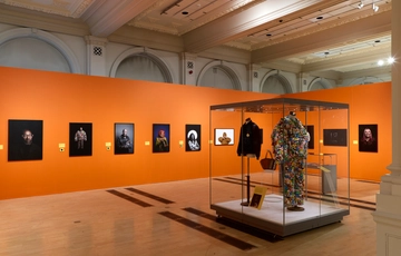 Gallery with orange walls displaying portrait photographs and a display case of two costumes.