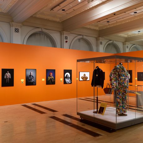 Gallery with orange walls displaying portrait photographs and a display case of two costumes.