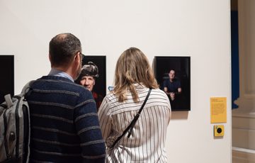 Two people observe framed portraits displayed on a white gallery wall.