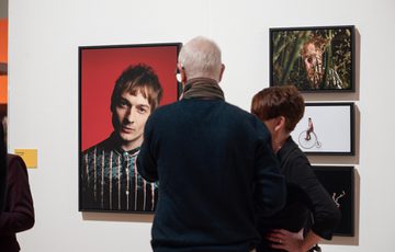 People observe framed portraits and artwork displayed on a white gallery wall.