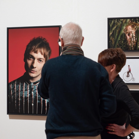 People observe framed portraits and artwork displayed on a white gallery wall.