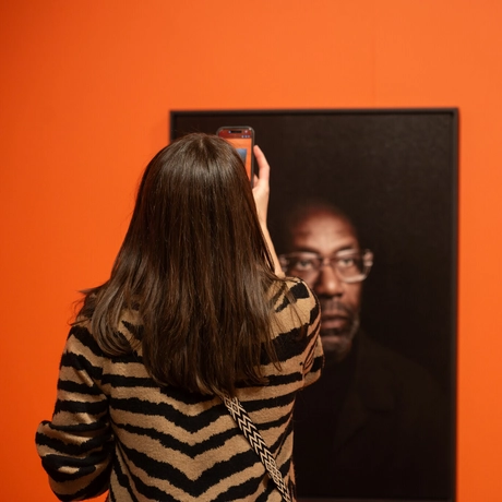 A woman captures a photo of a portrait of Lenny Henry on an orange museum wall.
