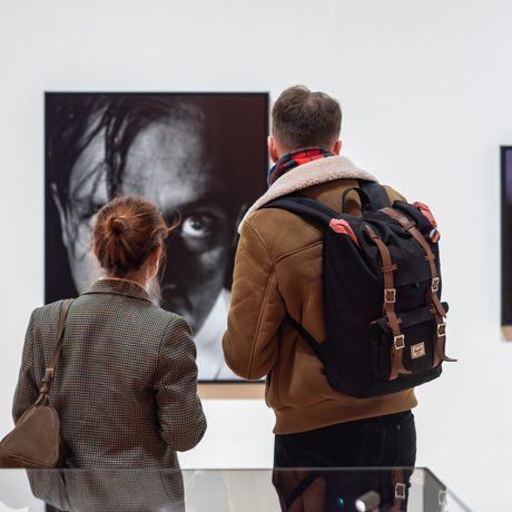 Two people closely observing large portrait artwork in a modern, white-walled gallery.