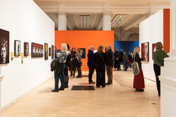 People observe and discuss portrait artwork displayed in a brightly coloured, spacious gallery.