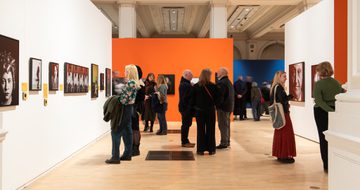People observe and discuss portrait artwork displayed in a brightly coloured, spacious gallery.