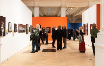 People observe and discuss portrait artwork displayed in a brightly coloured, spacious gallery.