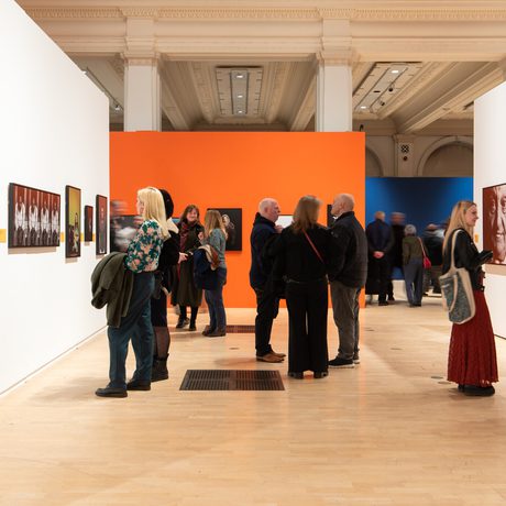 People observe and discuss portrait artwork displayed in a brightly coloured, spacious gallery.
