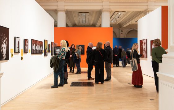 People observe and discuss portrait artwork displayed in a brightly coloured, spacious gallery.