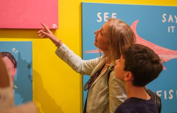 A woman and a boy observe colourful artwork on a bright yellow gallery wall.