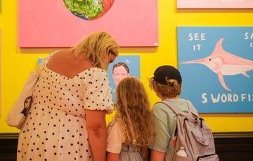 Woman and two children closely observing colourful paintings on a bright yellow wall.