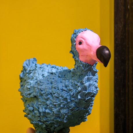 Sculpture of a blue dodo bird with a pink face and black beak on green grass.