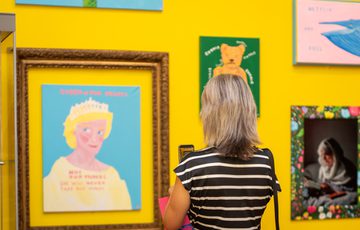 A woman in striped shirt views colourful framed artwork on a vibrant yellow wall.