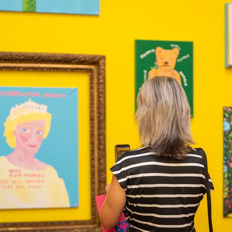 A woman in striped shirt views colourful framed artwork on a vibrant yellow wall.