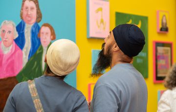 Two people observe colourful portraits displayed on vibrant yellow gallery walls.
