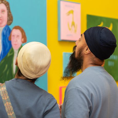 Two people observe colourful portraits displayed on vibrant yellow gallery walls.