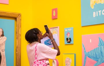 Person wearing pink captures colourful art pieces on bright yellow gallery walls with a smartphone.