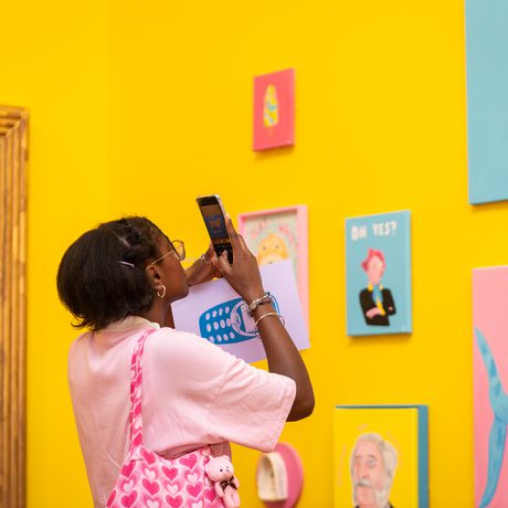 Person wearing pink captures colourful art pieces on bright yellow gallery walls with a smartphone.