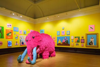 Bright yellow gallery walls decorated with colourful, animal and human paintings, featuring a large pink mammoth sculpture.