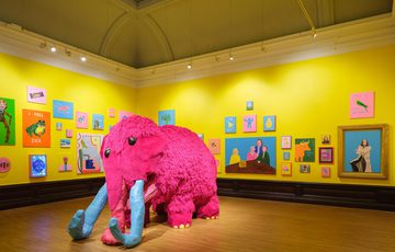 Bright yellow gallery walls decorated with colourful, animal and human paintings, featuring a large pink mammoth sculpture.