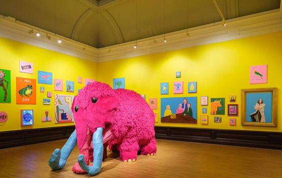 Bright yellow gallery walls decorated with colourful, animal and human paintings, featuring a large pink mammoth sculpture.