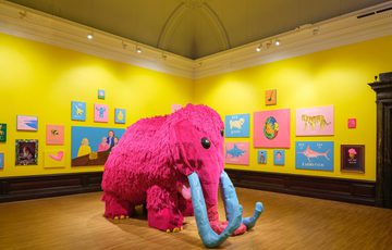 A large pink mammoth sculpture in the centre of a bright yellow gallery decorated with colourful paintings.