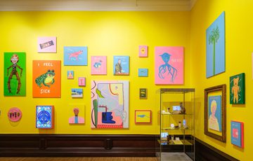 Colourful art pieces and display cabinet displayed against bright yellow walls in a gallery.