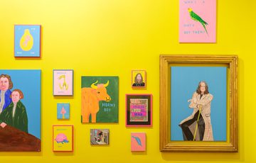 Colourful paintings displayed on a vibrant yellow wall in a gallery setting, one large painting is of Ozzy Osbourne in a large gold frrame..