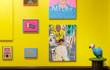 Colourful artworks on a bright yellow wall with a blue bird sculpture on a pedestal.