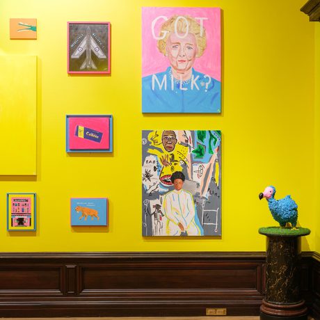 Colourful artworks on a bright yellow wall with a blue bird sculpture on a pedestal.