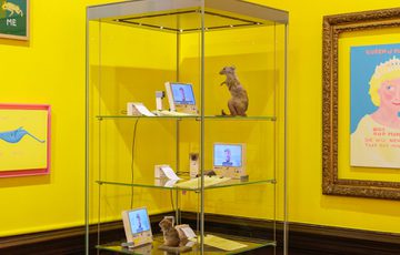 Glass display case with small sculptures and digital screens against bright yellow walls.