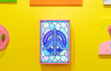 Colourful paintings stained glass displayed on a bright yellow wall alongside colourful paintings.