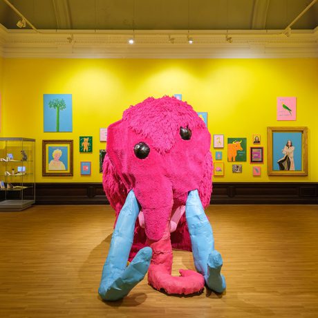 A large pink mammoth sculpture in the centre of a bright yellow gallery decorated with colourful paintings.