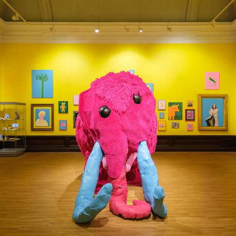 A large pink mammoth sculpture in the centre of a bright yellow gallery decorated with colourful paintings.