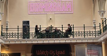 Lots of people in a gallery looking at a string quartet playing on the balcony above.
