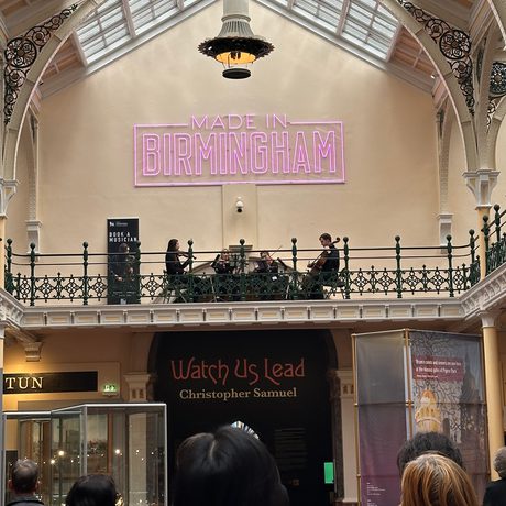 Lots of people in a gallery looking at a string quartet playing on the balcony above.