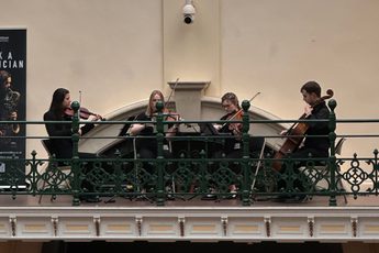 A string quartet playing on the balcony in a gallery, a pink neon 'Made In Birmingham' sign is above.