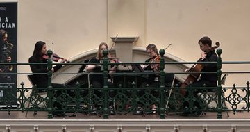 A string quartet playing on the balcony in a gallery, a pink neon 'Made In Birmingham' sign is above.