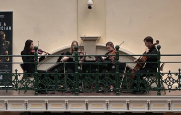 A string quartet playing on the balcony in a gallery, a pink neon 'Made In Birmingham' sign is above.