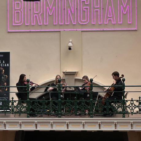 A string quartet playing on the balcony in a gallery, a pink neon 'Made In Birmingham' sign is above.