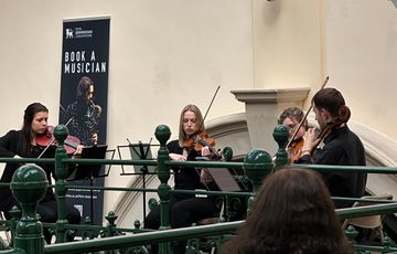 Side view of a string quartet playing on the balcony in a gallery, part of a pink neon sign can be seen above that reads 'Made In Birmingham'.