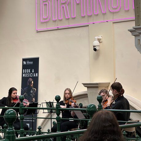 Side view of a string quartet playing on the balcony in a gallery, part of a pink neon sign can be seen above that reads 'Made In Birmingham'.