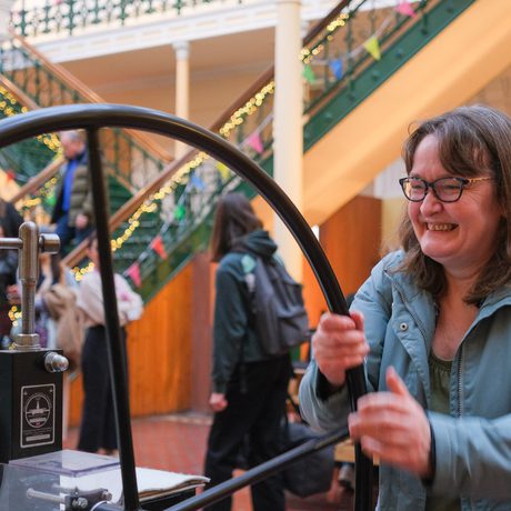 A person is smiling and enthusiastically operates a large hand wheel of a printing press.