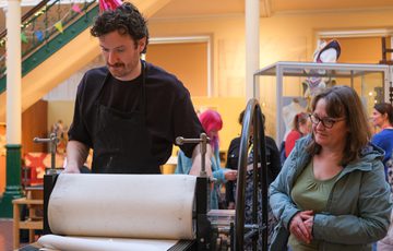 A person operates printing press while another person watches.