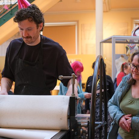 A person operates printing press while another person watches.