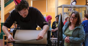 Person wearing prints on a press while others watch attentively.