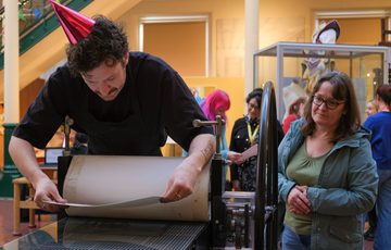Person wearing prints on a press while others watch attentively.