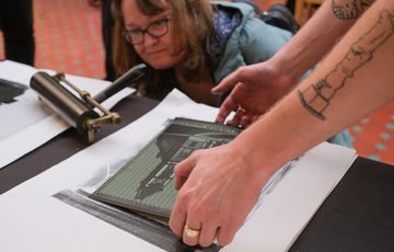 Person carefully placing a printing plate while another observes closely during printmaking process.