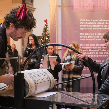 Two people operate a printing press while onlookers observe.